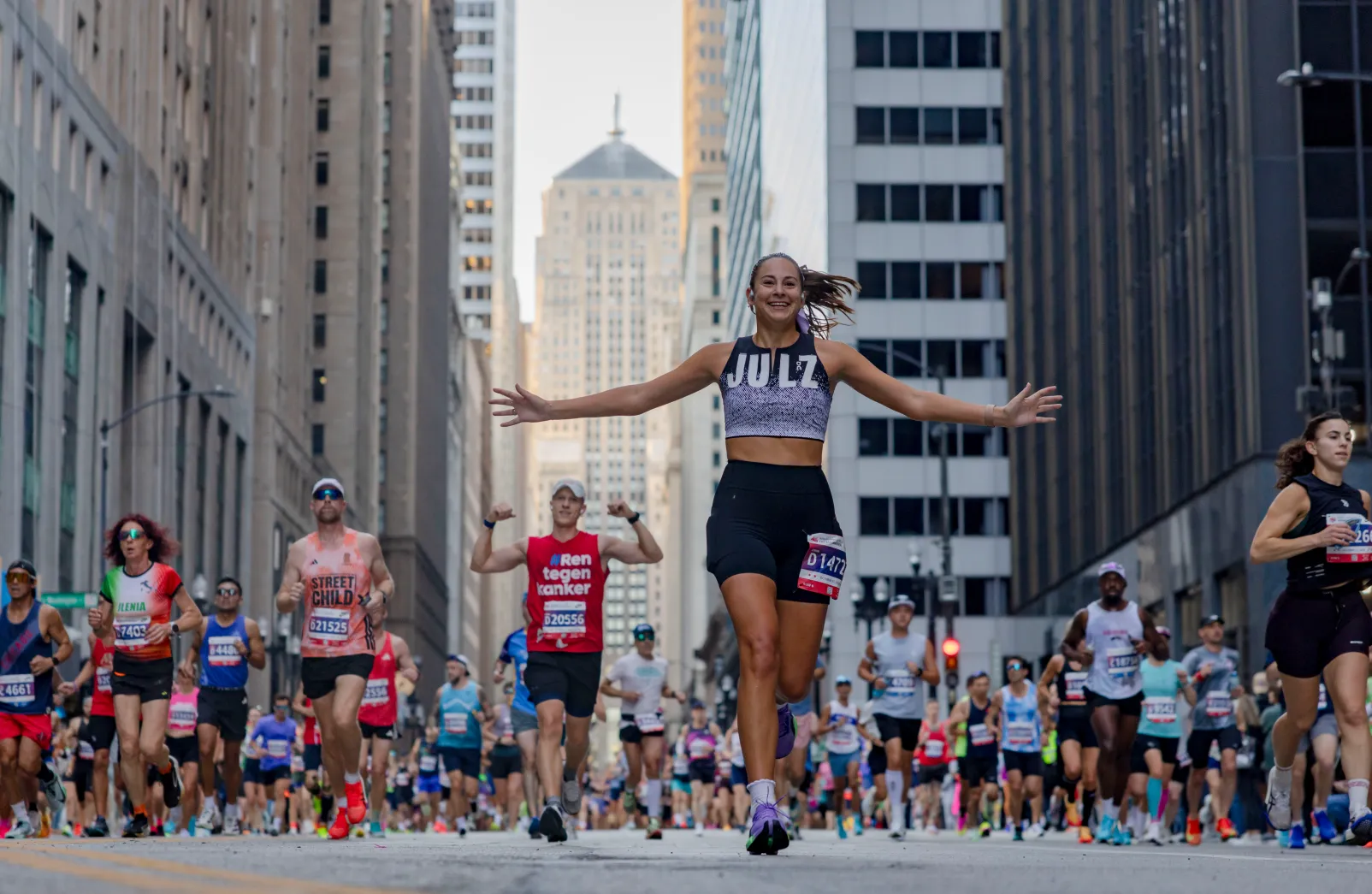 A smiling runner with arms spread wide leads a pack of marathoners between downtown towers