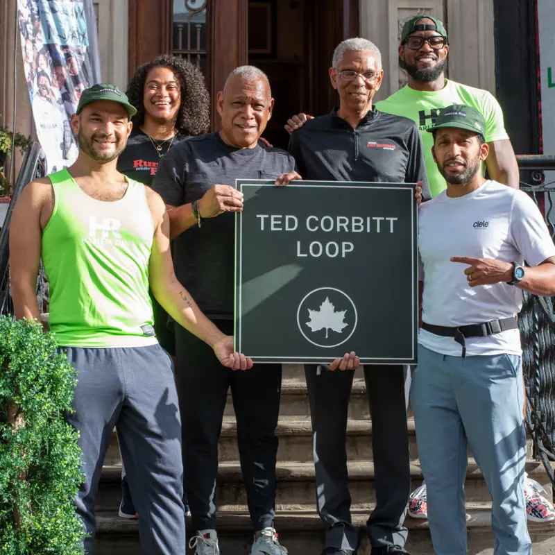 Runners holding up a Ted Corbitt Loop sign on brownstone steps